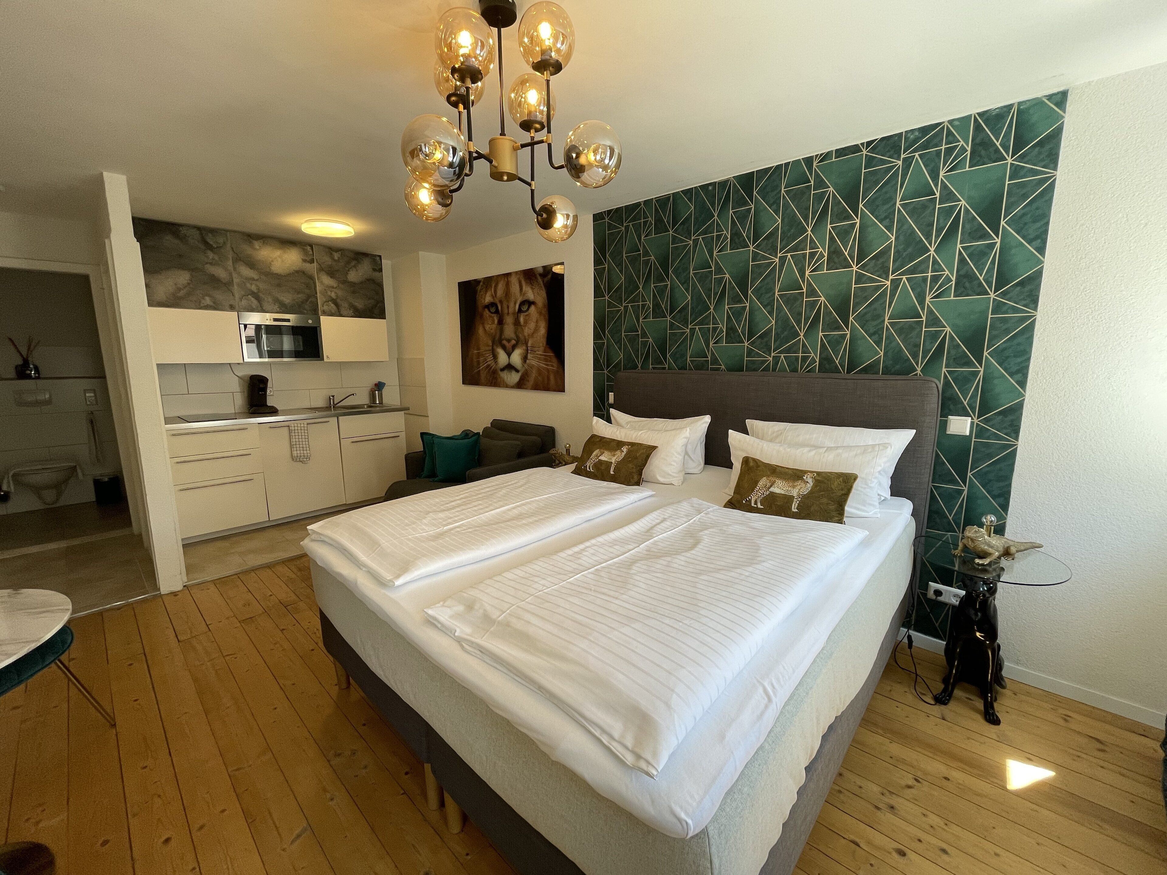 Ett modernt hotellrum med en stor säng, ett litet kök, ett badrum och en unik väggdesign med geometriska mönster.