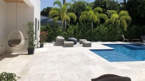 Un espace piscine extérieur moderne, doté d’un patio carrelé, de chaises longues et d’un fauteuil suspendu.