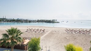 Een strand met parasols en ligbedden, een palmboom en uitzicht op de zee en gebouwen in de verte.