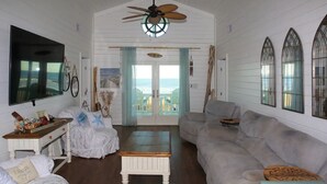 En rummelig stue med en stor grå sofa, et hvidt sofabord og en loftsvifte. Der er et stort vindue med havudsigt, og en dør til en balkon.