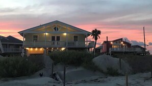 Et hus ved stranden med balkon og en ekstra etage, omgivet af sand og beplantning.