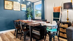 A dining area with a wooden table, chairs, and a baby high chair. There are framed botanical prints on the wall, a window with curtains, and a lamp on a side table. - The Jungalow: 65" 4K TV, memory foam, kid-friendly (Louisville)