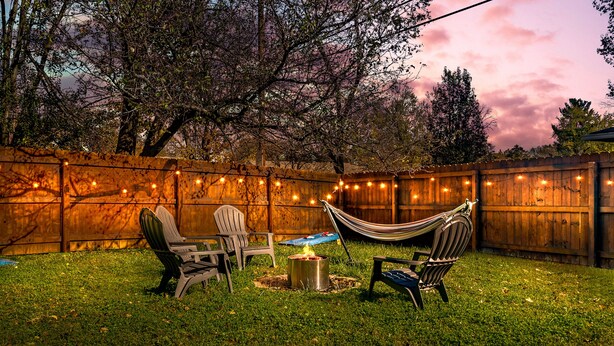 A backyard with a wooden fence, string lights, a fire pit, and two chairs. - The Jungalow: 65" 4K TV, memory foam, kid-friendly (Louisville)