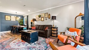 A modern living room with a sofa, ottoman, and armchair, a dining area with chairs and a table, and a large rug. - The Jungalow: 65" 4K TV, memory foam, kid-friendly (Louisville)