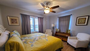 Ein Schlafzimmer mit einem Bett, zwei Stühlen, einem Deckenventilator, einem Nachttisch und einem Fenster mit Vorhängen.