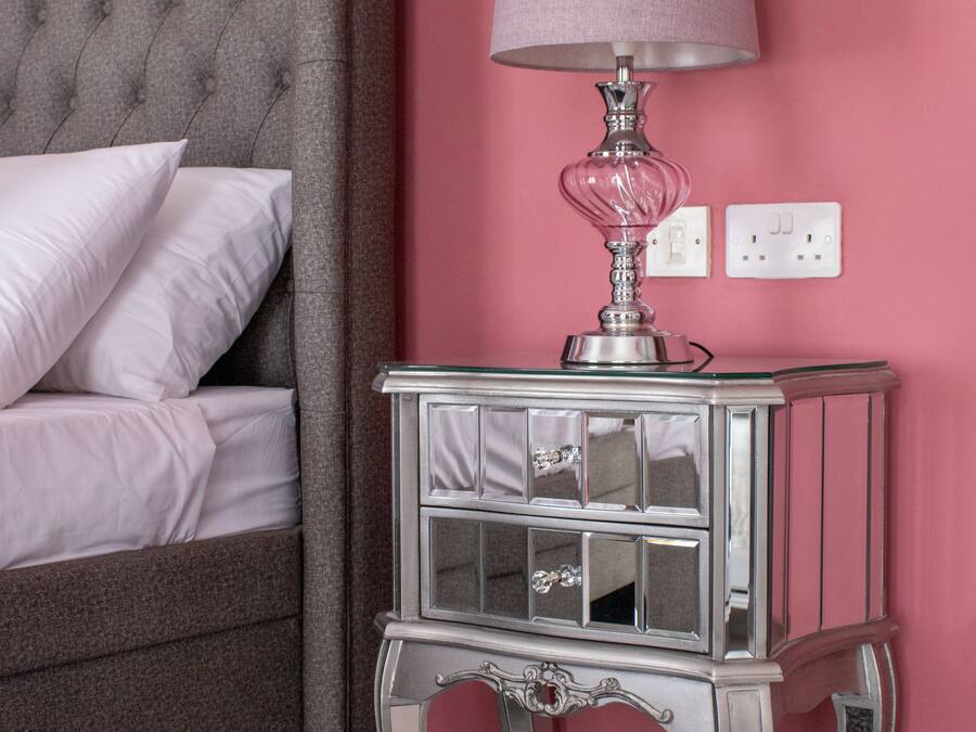 Un dormitorio con cabecera capitonada, una mesita de noche con lámpara y pared rosa.