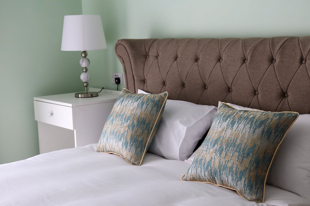 Un lit bien fait, avec du linge de lit blanc et deux coussins décoratifs, une table de chevet avec une lampe et une prise électrique au mur.