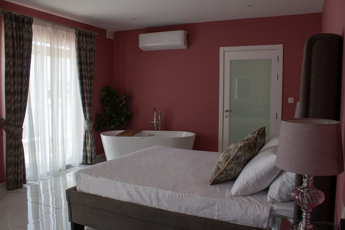 Une chambre à coucher avec un lit, une lampe de chevet, une salle de bain avec une baignoire et une fenêtre avec des rideaux.