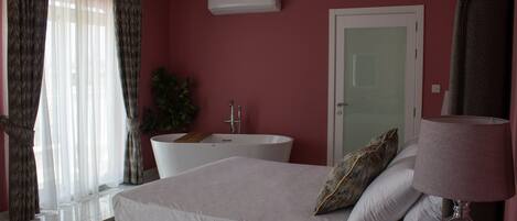 Une chambre à coucher avec un lit, une lampe de chevet, une salle de bain avec une baignoire et une fenêtre avec des rideaux.