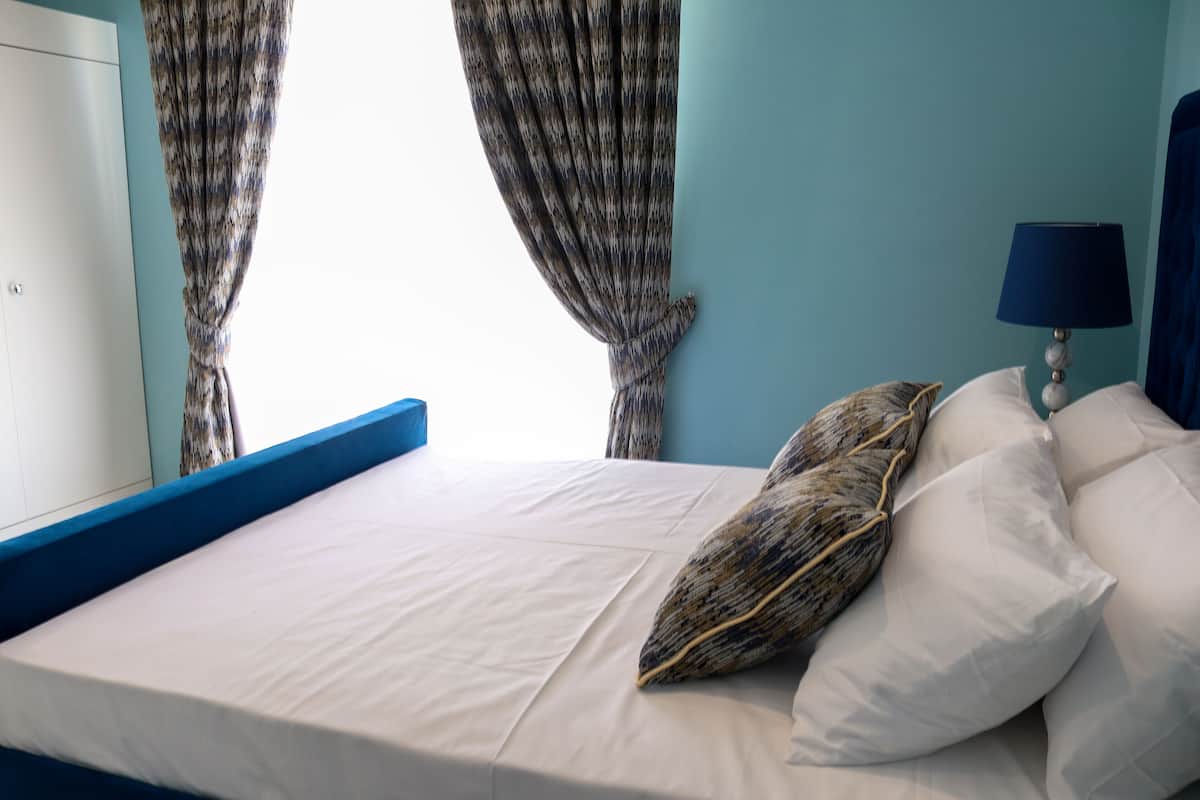 Un lit bien fait, avec du linge de lit blanc et un coussin à motifs, une lampe de chevet et une vue sur une fenêtre à travers les rideaux.