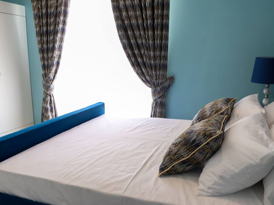 Un lit bien fait, avec du linge de lit blanc et un coussin à motifs, une lampe de chevet et une vue sur une fenêtre à travers les rideaux.