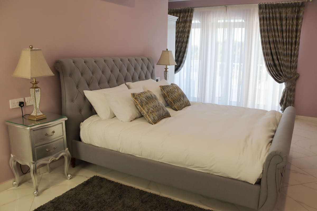 Un lit bien fait, avec du linge de lit blanc et deux coussins décoratifs, une table de chevet avec une lampe et une fenêtre ornée de rideaux légers.