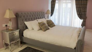 Uma cama bem arrumada com lençóis brancos e duas almofadas decorativas, uma mesa de cabeceira com abajur e uma janela com cortinas leves.