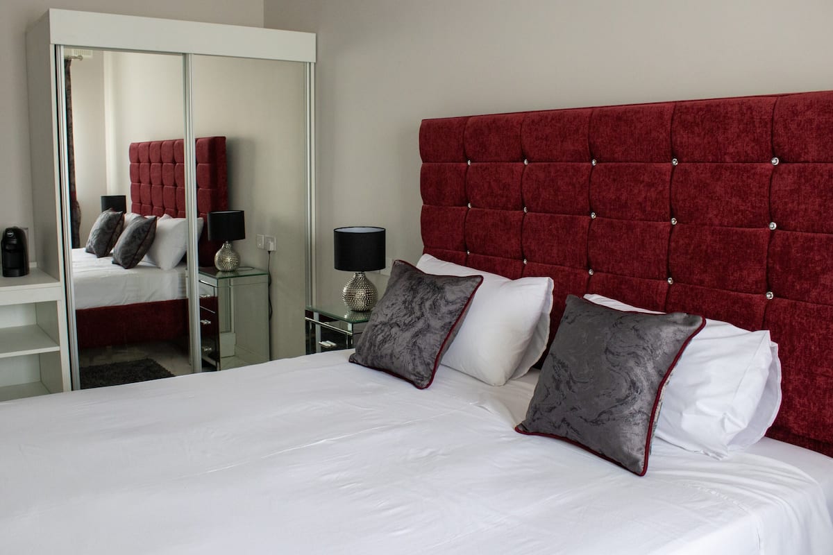 Un lit bien fait, avec du linge de lit blanc et deux oreillers gris, une tête de lit rembourrée en tissu rouge, une table de chevet avec une lampe et un miroir accroché au mur.