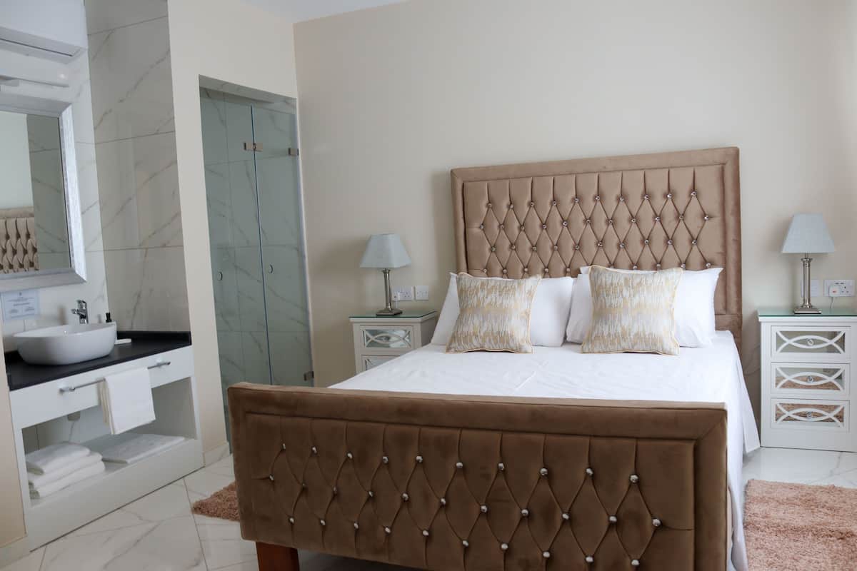 Une chambre à coucher avec une tête de lit capitonnée, un grand lit, deux tables de chevet et une salle de bain équipée d’un lavabo et d’un miroir.
