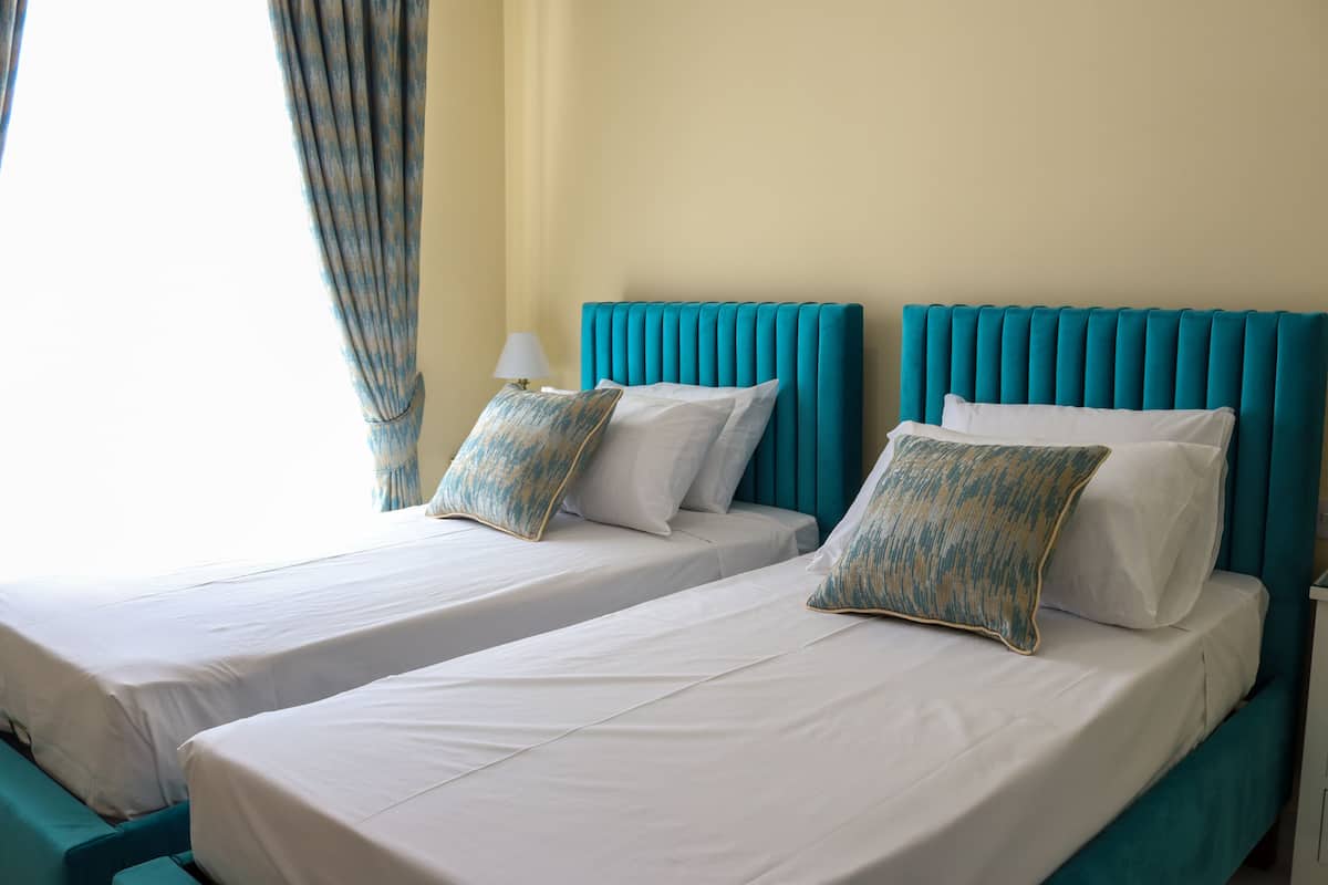 Deux lits jumeaux avec du linge de lit blanc et des têtes de lit couleur turquoise, une lampe et une fenêtre avec des rideaux.