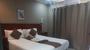 A hotel room with a neatly made bed, a nightstand, a wall-mounted lamp, and a window with curtains. - ShakeVille Guest House (Shakawe)