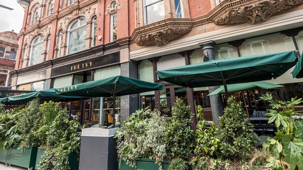 En historisk byggnad med utsmyckad arkitektur, en butik som heter ”The Ivy” med gröna markiser och frodig grönska framför.