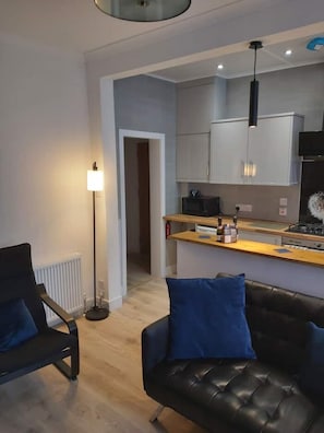 A modern living room with a black leather sofa and a blue armchair, a kitchen with white cabinets and a black stove, and a floor lamp. - Apartment49Oban, very close to Town center. (Oban)