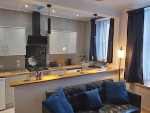 A modern kitchen with white cabinets, a black stove, and a wooden countertop. There is a black leather sofa with blue cushions in the foreground. - Apartment49Oban, very close to Town center. (Oban)