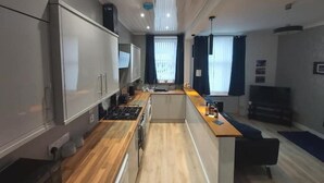 Moderni keittiö, jossa on keskeinen saareke, integroidut kodinkoneet sekä ruokailutila, jossa on tumma sohva ja televisio.