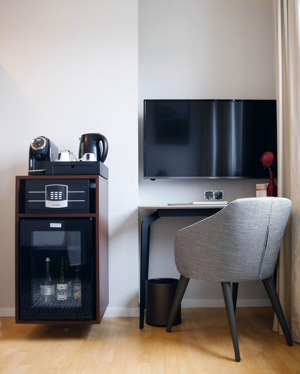 A hotel room with a desk, a chair, a television, a minibar, and a trash bin. - Villa Bosco - Boutiquehotel (Walldorf)