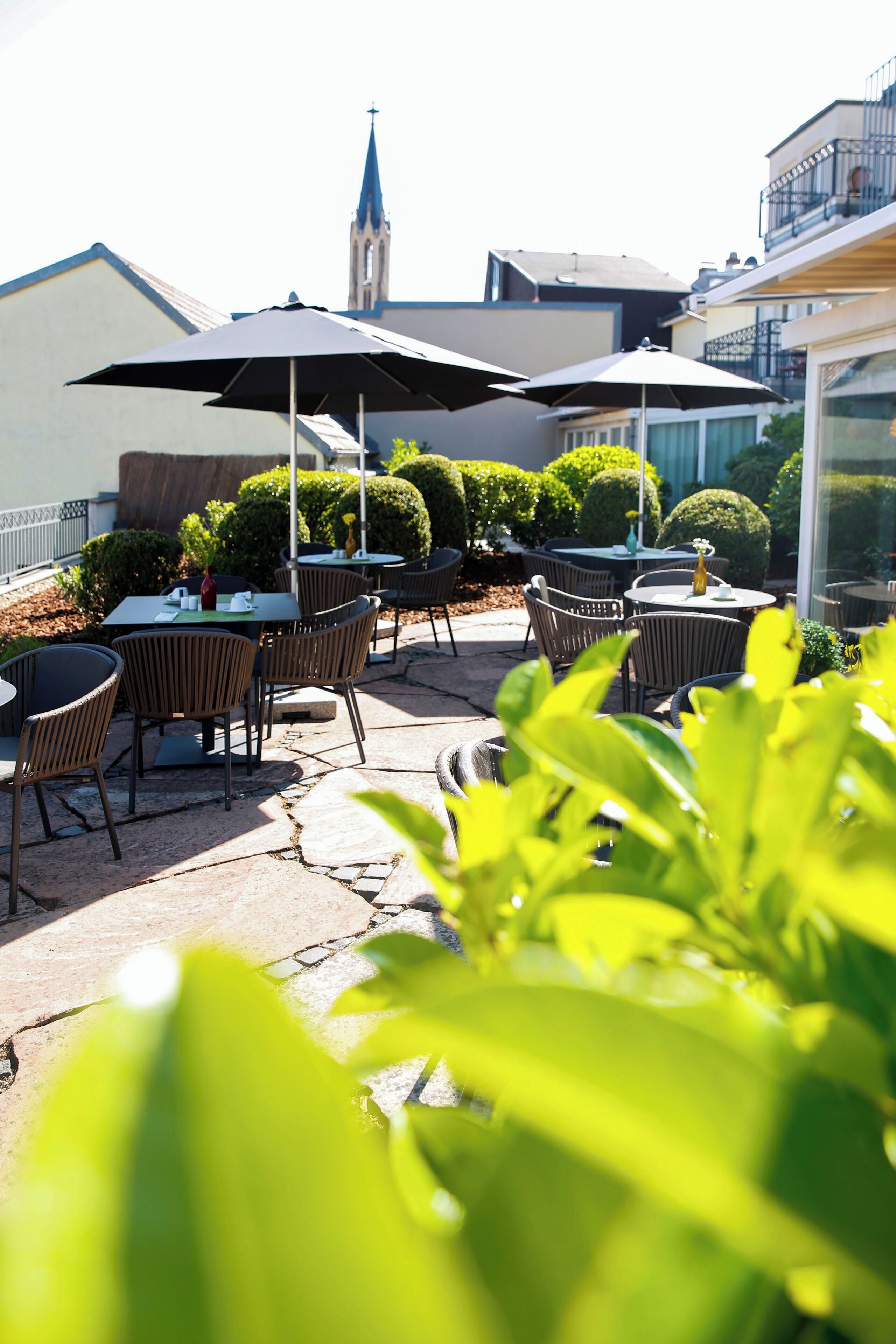 An outdoor patio with tables and chairs, surrounded by greenery and a building with a spire in the background.