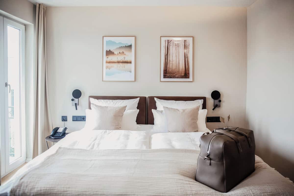 Una camera d'albergo con un letto grande, due quadri appesi al muro, un comodino con un telefono e una valigia sul letto.