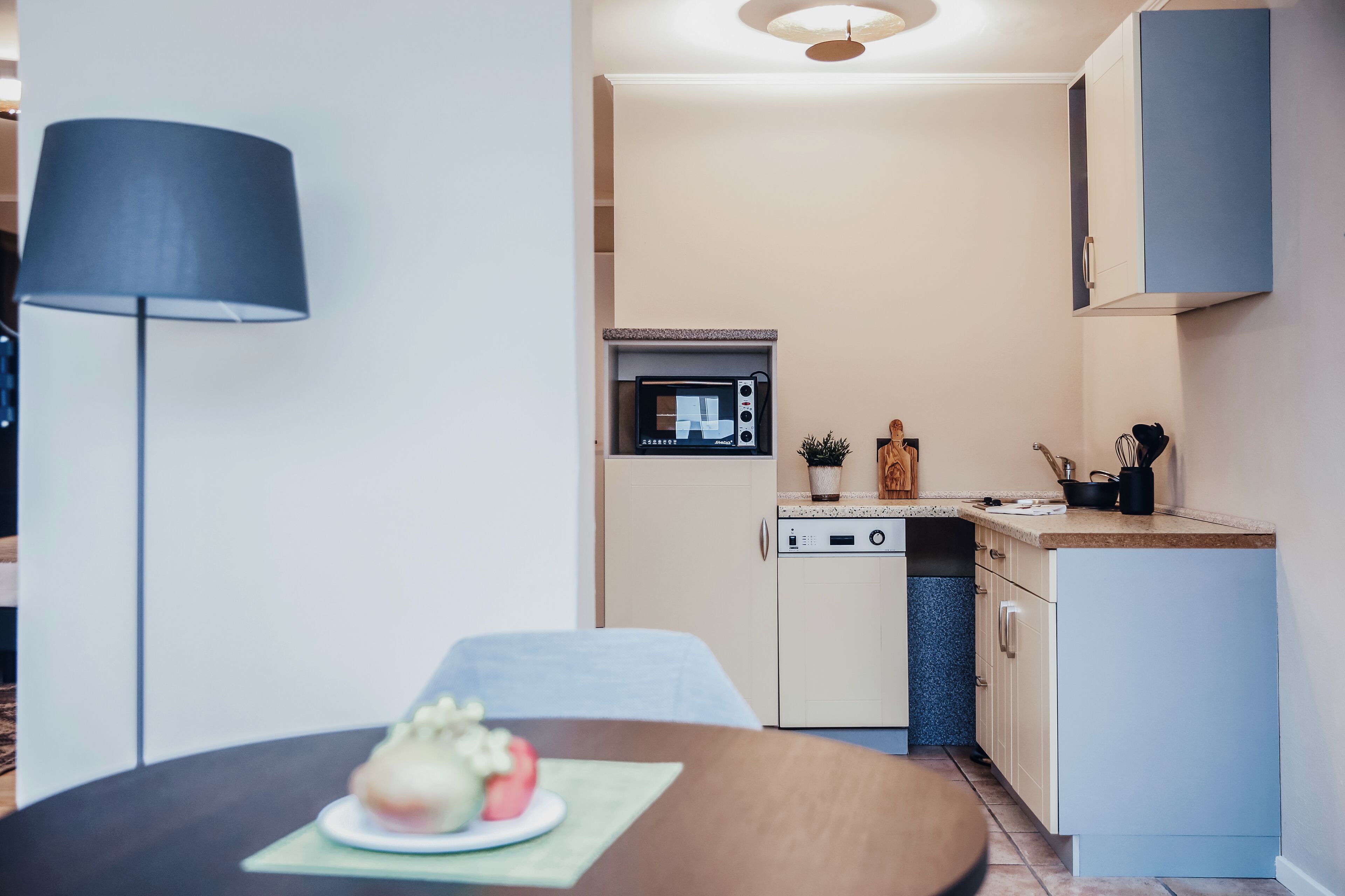 Een praktische keuken met een magnetron, oven en spoelbak. Op de voorgrond staat een eettafel met een bord eten en een lamp.