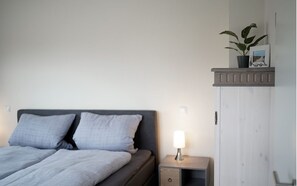 A bedroom with a bed, bedside table, nightstand, and a shelf with a plant and picture frame.