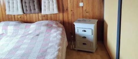 A bedroom with a wooden ceiling, a bed with a checkered blanket, a nightstand with drawers, and a wall-mounted shelf with pillows.