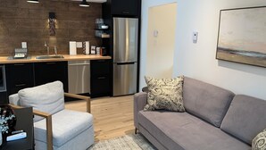 Una sala de estar moderna con un sofá gris, un sillón gris y una mesa de centro de madera. Hay una zona de cocina con encimera negra, electrodomésticos de acero inoxidable e iluminación colgante.