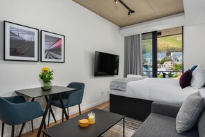 A modern hotel room with a bed, a TV, a dining table with chairs, and a view of buildings through large windows.