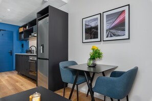 A modern kitchen with a blue door, a built-in oven, and a refrigerator. There is a dining area with a table and two chairs, and a framed picture on the wall.