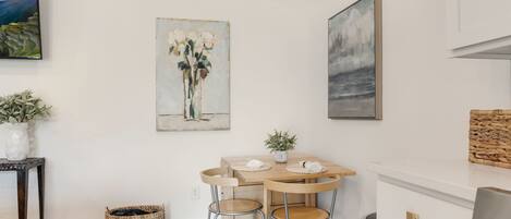 A modern kitchen with a wooden dining table, two chairs, and a small potted plant. There is a woven basket on the floor and a framed painting on the wall.