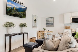 A modern living room with a sofa, a coffee table, and a TV mounted on the wall. There is a wooden desk with a chair and a small plant on it. -  Adorable Upscale cottage in Downtown Fountain Inn.  (Fountain Inn)