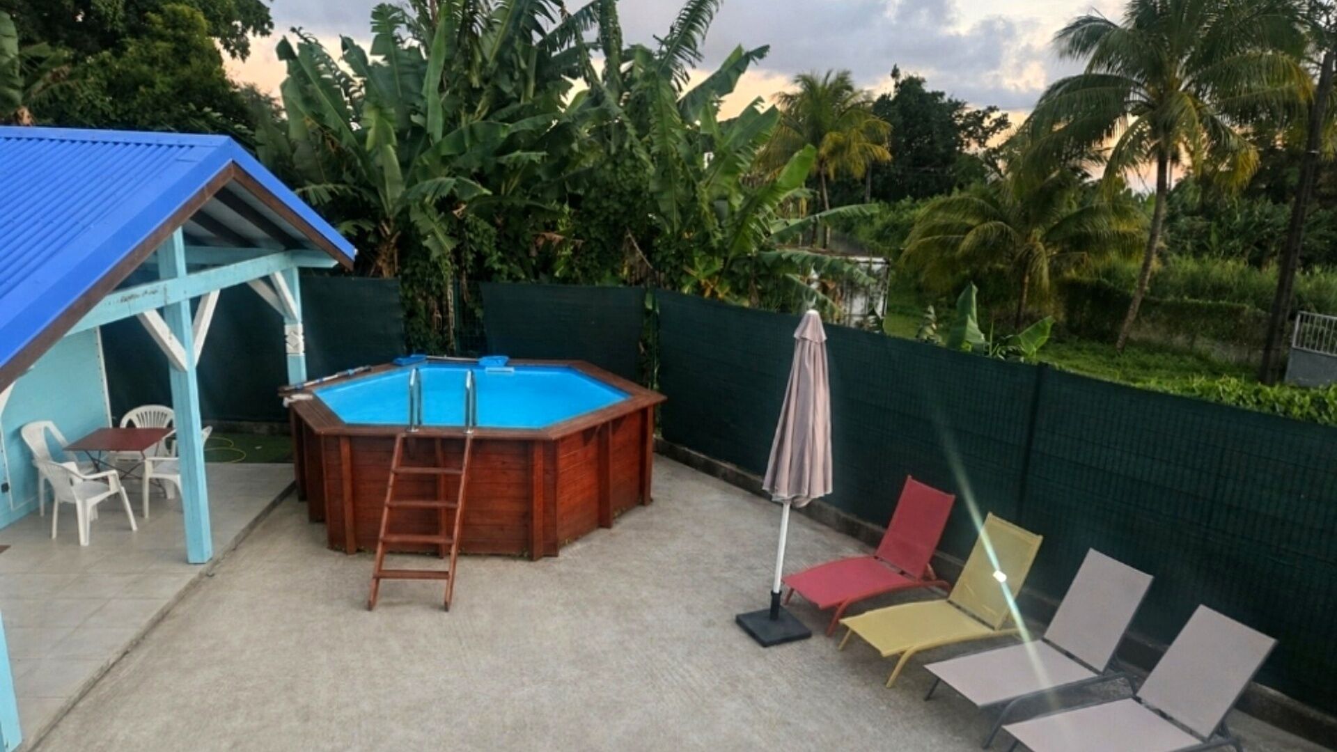 Una piscina exterior amb una terrassa de fusta, una zona de seients coberta i vista a vegetació tropical.