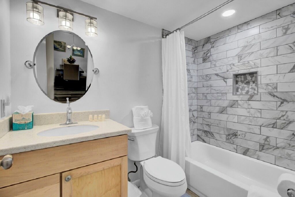 A bathroom with a marble-tiled shower wall, a white toilet, a sink with a mirror above it, and wooden cabinets.