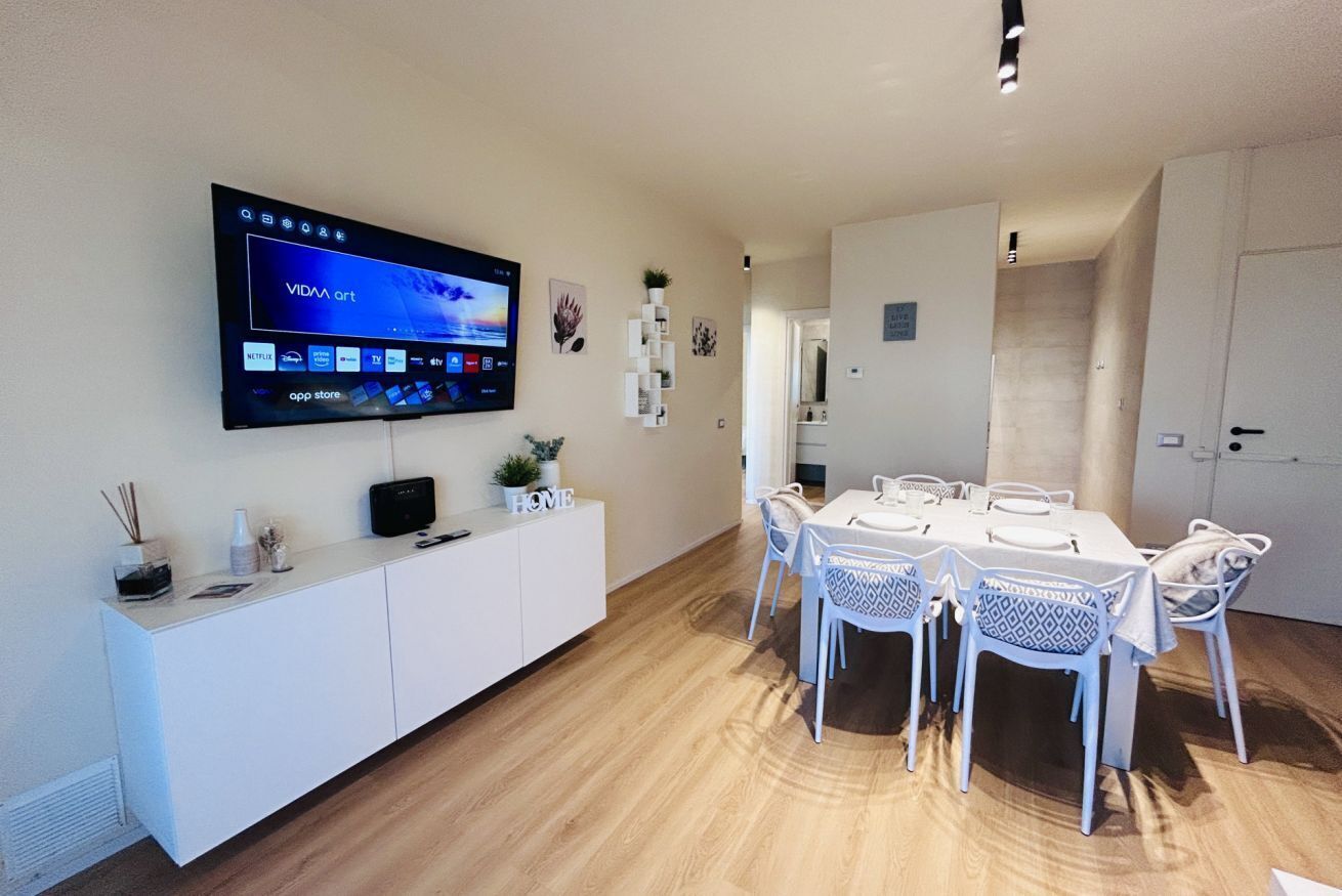 Ett modernt matområde med en tv monterad på väggen som visar konst från ”VIDA”. Här finns ett vitt konsolbord med dekorativa föremål och ett vitt matbord med mönstrade dukar och stolar.