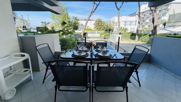 Uma zona de refeições com uma mesa posta para quatro pessoas, rodeada de cadeiras, num pátio pavimentado com vista para edifícios e árvores.