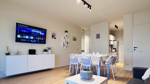 Uma sala de estar moderna com uma TV de tela plana, móveis brancos e um espaço de refeições com cadeiras e uma mesa.