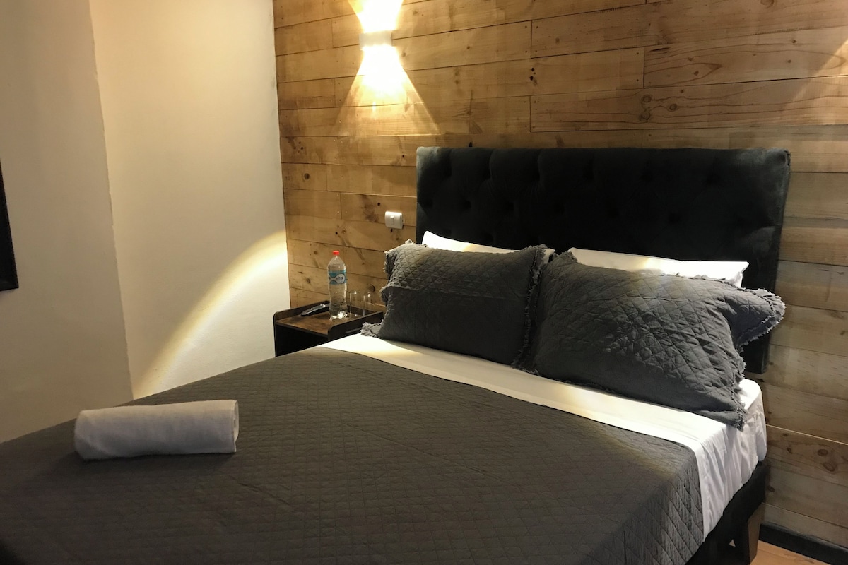 A bed with a quilted grey bedspread, two grey pillows, and a white pillow. There is a water bottle and a remote control on a wooden nightstand beside the bed.