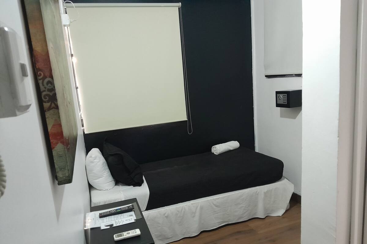 Une petite chambre avec un lit simple, un matelas noir, des oreillers blancs, une petite table de chevet avec une télécommande et un rideau blanc.