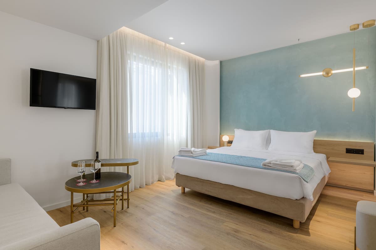 A modern hotel room with a bed, a TV, a small round table with a bottle and glasses, and a large window with curtains.