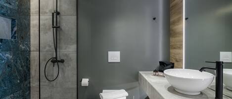 A modern bathroom with a large white sink, a black faucet, a shower with a black hose, a white toilet, and a trash can.