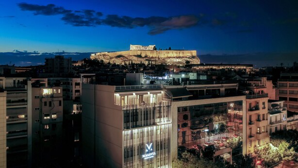 A cityscape with a prominent ancient structure on a hill, modern buildings, and a well-lit commercial building with a visible logo. - Acropolis Magenta (Athens)