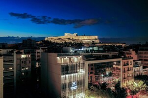 A cityscape with a prominent ancient structure on a hill, modern buildings, and a well-lit commercial building with a visible logo.