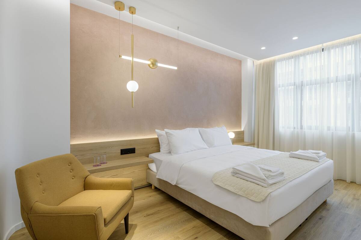 A modern hotel room with a large bed, a bedside table, a chair, and a large window with sheer curtains.