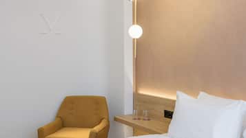 A modern hotel room with a minimalist design, featuring a single armchair, a bedside table with two glasses, and a wall-mounted light fixture.