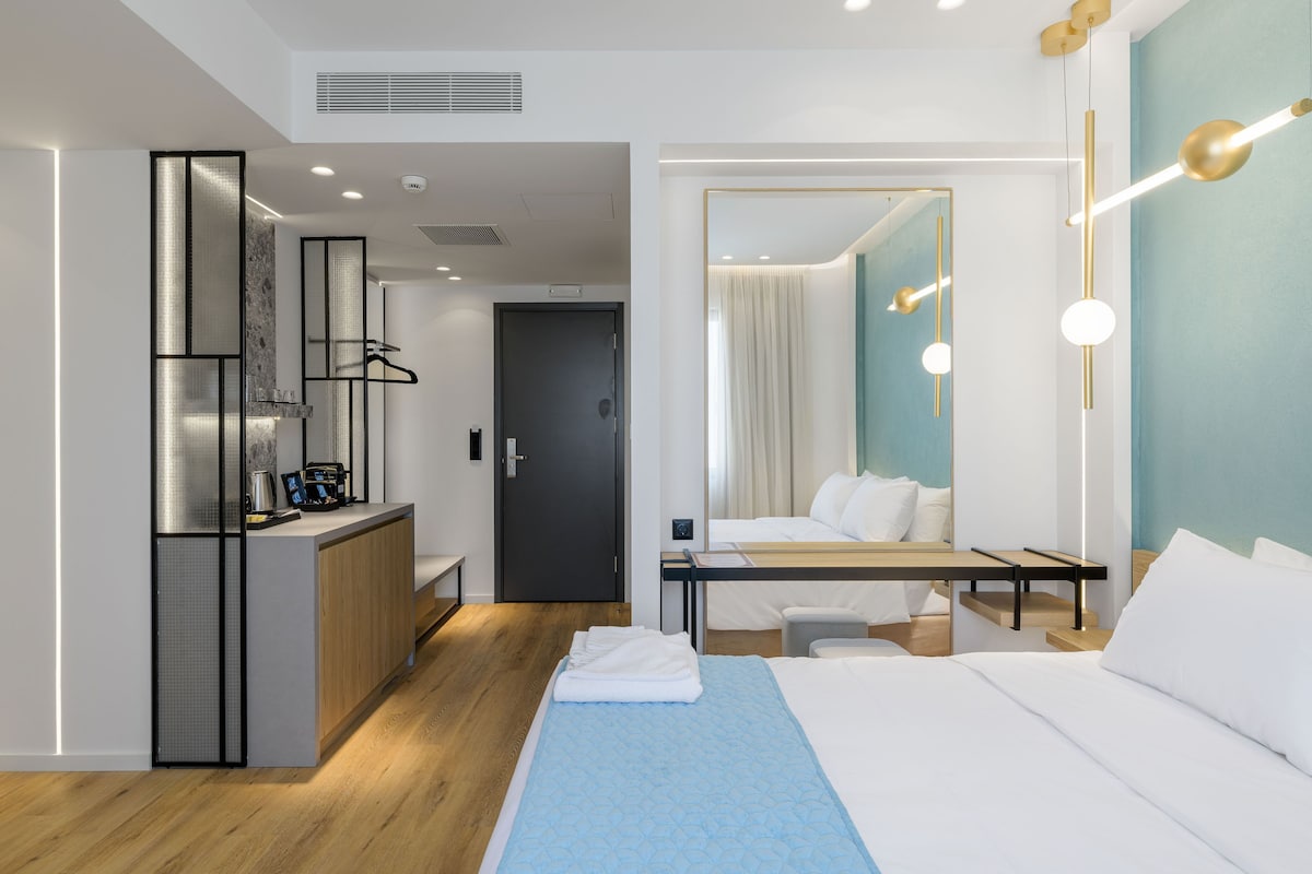 A modern hotel room with a bed, a desk, a mirror, and a wardrobe.